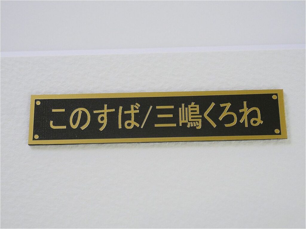 Gold-bordered black plaque mounted on a white wall, displaying Japanese text: "このすぱ/三嶋くろね".